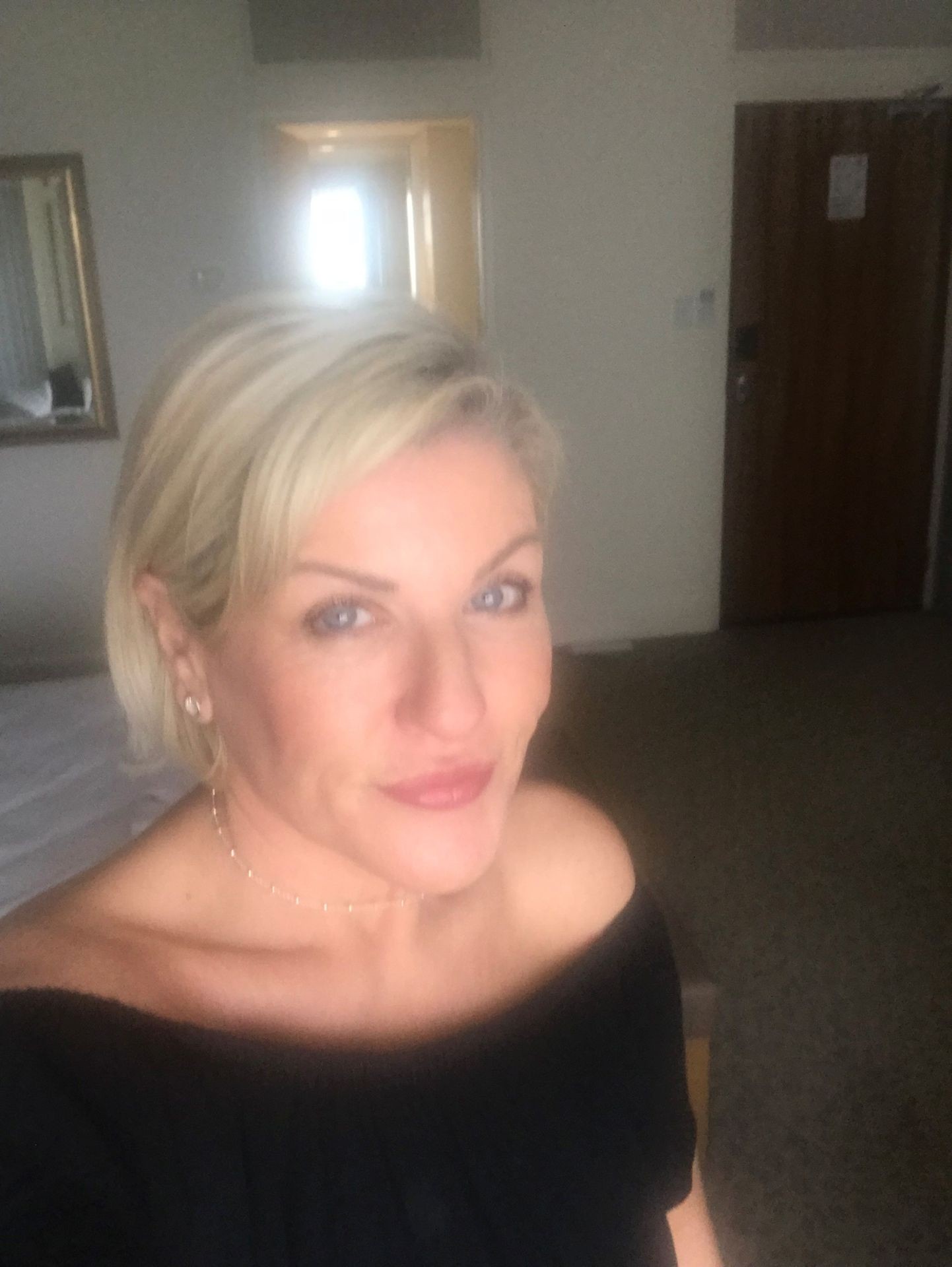 Person with short blonde hair in a room with a bed and a door in the background.